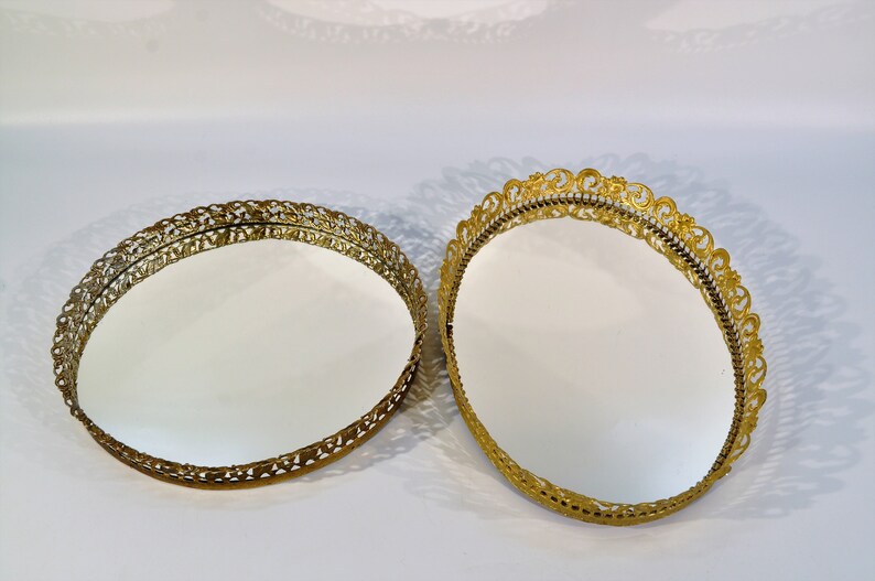 Puede incluir: Dos bandejas redondas decorativas de color dorado con bordes adornados con filigranas. Las bandejas tienen una superficie de espejo.