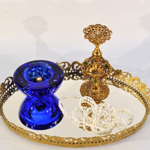 Puede incluir: Una bandeja de espejo dorado con un borde decorativo de filigrana. La bandeja presenta un cuenco de vidrio azul, un frasco de perfume dorado con joyas de colores y un collar de perlas.