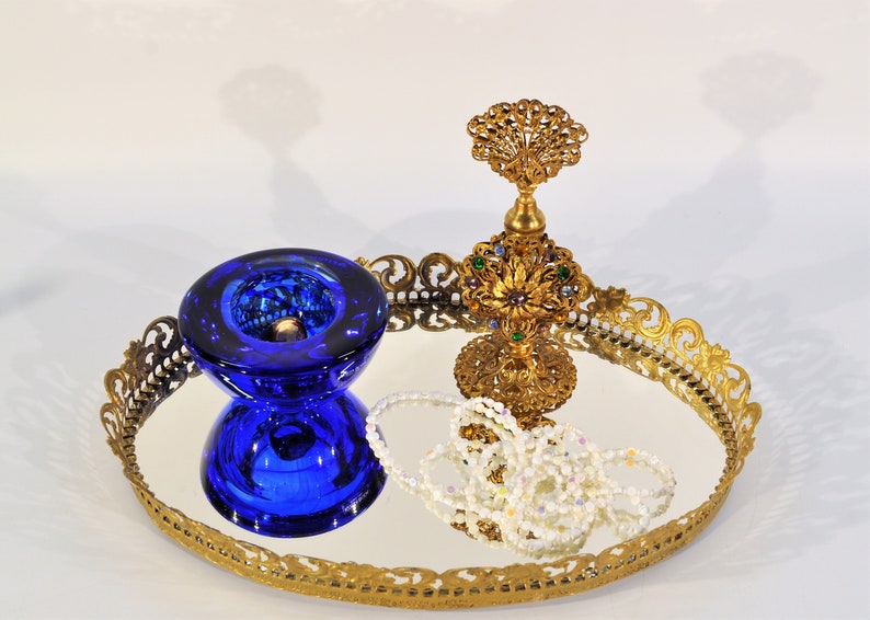 Puede incluir: Una bandeja de espejo dorado con un borde decorativo de filigrana. La bandeja presenta un cuenco de vidrio azul, un frasco de perfume dorado con joyas de colores y un collar de perlas.