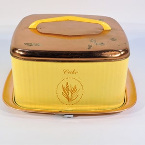 May include: A yellow and copper cake carrier with a handle. The carrier has a wheat design and the word "Cake" on the front.