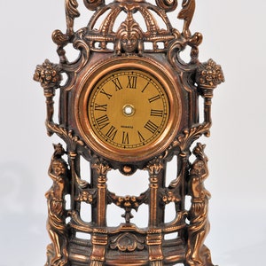 May include: A gold-toned, ornate clock with a Roman numeral face and a quartz movement. The clock is on a decorative stand with two figures on either side.