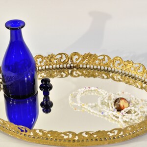 Puede incluir: Una bandeja de espejo ovalada adornada con un marco dorado. Una botella de vidrio azul y un peque&ntilde;o tap&oacute;n de vidrio azul est&aacute;n en la superficie de la bandeja. Un collar de cuentas blancas y multicolores tambi&eacute;n est&aacute; en la bandeja.