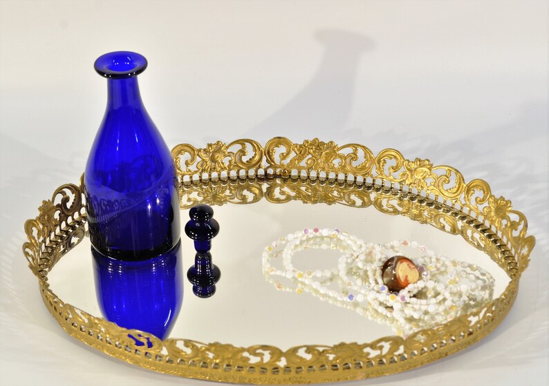Puede incluir: Una bandeja de espejo ovalada adornada con un marco dorado. Una botella de vidrio azul y un peque&ntilde;o tap&oacute;n de vidrio azul est&aacute;n en la superficie de la bandeja. Un collar de cuentas blancas y multicolores tambi&eacute;n est&aacute; en la bandeja.