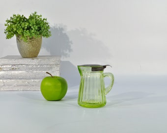 May include: A green glass pitcher with a metal lid and a ribbed design. The pitcher is sitting on a white surface with a green apple and a potted plant in the background.