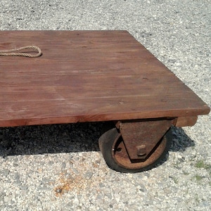 May include: A rectangular wooden industrial cart with a dark brown finish, featuring a thick rope handle attached to one side. The cart is supported by two large, heavy-duty rusted metal wheels on one end and two metal legs on the other end.