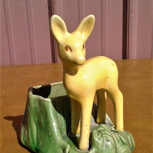 May include: A vintage ceramic planter featuring a yellow deer figurine standing in a green planter. The deer has large ears and a gentle expression. The planter has a textured, leaf-like design.