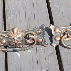 May include: Two antique bronze drawer pulls with ornate floral and scroll designs. Each pull has a curved handle and is mounted on a rectangular backplate with decorative accents. The pulls are set against a weathered wooden surface.
