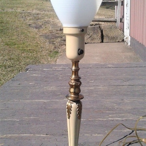 May include: An antique table lamp with a white, cone-shaped shade. The lamp features a cream-colored base and stem with ornate gold-colored accents. The base is round with decorative feet. A power cord extends from the base.