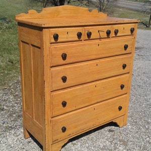 May include: A tall, antique wooden dresser with six drawers and a decorative top. The dresser is painted a light yellow color with dark brown knobs. The keyhole is visible on the top drawer. The dresser is standing on a gravel surface.