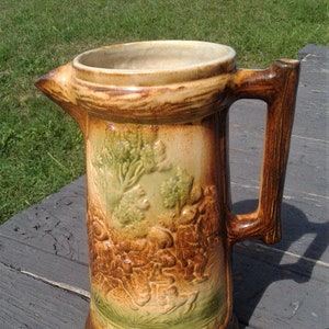 May include: A ceramic pitcher featuring a raised relief design of a forest scene with figures and a dog. The vessel has a glossy brown and tan glaze, a wide circular rim, and a sturdy handle shaped like a textured tree branch or piece of wood.