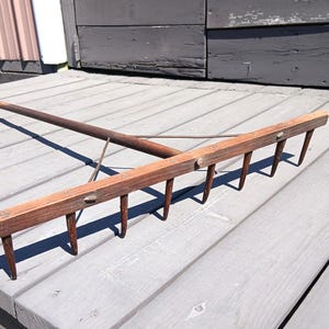 May include: A wooden garden rake with a long, brown handle and a head with several metal tines. The rake is made of wood and metal, and it is designed for gardening tasks. The rake is lying on a gray wooden surface.