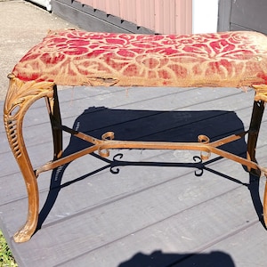 May include: An antique bench with a red floral patterned cushion and ornate, carved wooden legs. The legs are a warm, golden brown color, and the bench has a metal support bar. The bench is placed on a wooden deck.