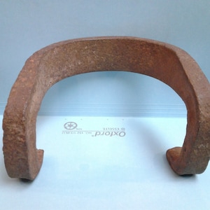 May include: A weathered, U-shaped metal object with a rusty brown color. The object has a rough, textured surface and appears to be made of iron. The shape suggests it may be a horseshoe or a similar agricultural tool.