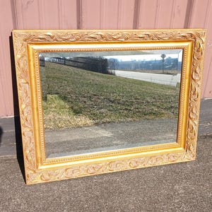 May include: A rectangular wall mirror featuring a wide, ornate gold-colored frame with intricate carved leaf and vine patterns. The inner edge of the frame displays a delicate beaded texture, surrounding a clear, reflective glass surface.