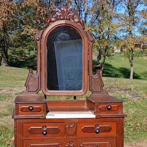 May include: A vintage wooden dresser with a large mirror. The dresser has multiple drawers with dark knobs and decorative carvings. The mirror is framed and has a decorative top. The wood has a warm, reddish-brown tone.