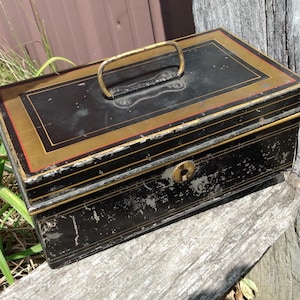 May include: A rectangular black metal lockbox featuring a gold-colored metal handle on the lid and a central keyhole on the front. The box has decorative gold and red painted border lines on the lid and shows signs of paint wear and aging.
