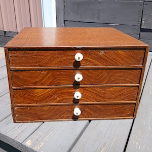 May include: A rectangular wooden storage chest featuring four horizontal drawers stacked vertically. Each drawer is finished with a prominent wood grain pattern and includes a single small, round, white knob centered on the front panel.