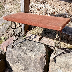 May include: A small, rectangular bench with a reddish-brown top and ornate, dark metal legs. The bench is supported by decorative, curved metalwork and a horizontal bar. The bench is outdoors, resting on large stones.