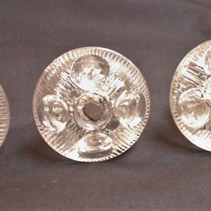 May include: Four clear glass cabinet knobs with a circular design. Each knob features a central dark circle surrounded by four smaller circles and a textured, ridged pattern. The knobs are arranged in a row against a dark background.