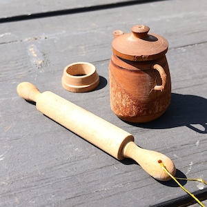 May include: A light-colored wooden rolling pin with a thin gold string attached to one handle, a small wooden ring, and a dark brown wooden barrel-shaped container with a lid and a small handle, all arranged together on a flat surface.