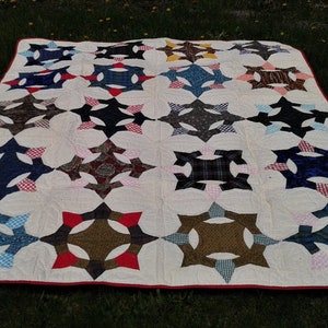 May include: A rectangular quilt featuring a repeating star and circle pattern in various plaid and solid fabrics. The design is set against a cream-colored background with a thin red border edge, showcasing a mix of blue, brown, and black tones.