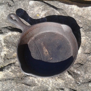 May include: A small, well-worn cast iron skillet with a single handle. The pan is dark brown with a slightly textured surface, showing signs of use. The handle has a loop for hanging. The skillet is sitting on a rough, gray stone surface.