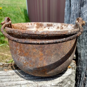 May include: A small, antique cast iron cauldron with a rusty brown finish. The cauldron has a rounded body, a wide rim, and two arched handles. It sits on a weathered wooden surface, with a blurred green background.