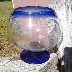 May include: A clear glass bowl with a rounded body and a cobalt blue base and rim. The bowl is sitting on a weathered wooden surface. The glass reflects light, creating bright spots.