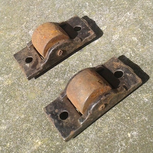 May include: Two vintage metal rollers with a rectangular base and a rounded, cylindrical roller. The metal has a rusty brown color on the roller and a dark, aged appearance on the base. Each base has two holes for mounting.