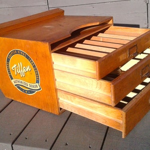 May include: A vintage wooden storage cabinet with three pull-out drawers featuring internal dividers. The side of the cabinet displays a circular yellow and black Tiffen Authorized Dealer logo with text reading Filters, Adapter Rings, Lens Shades.