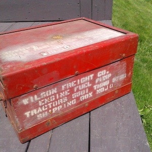 May include: A weathered red metal shipping box with the text "WILSON FREIGHT CO. ENGINE FUEL PUMP TRACTORS 5501 THRU 8786 SHIPPING BOX NO.10" in white lettering. The box is rectangular and sits on a wooden surface.