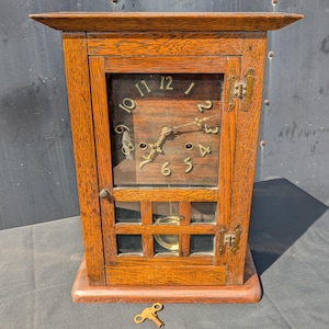 May include: A rectangular wooden mantel clock with a dark brown finish, featuring a glass-fronted door, brass hinges, and a brass winding key. The clock face displays gold-colored Arabic numerals and ornate hands, with a visible pendulum behind a grid.