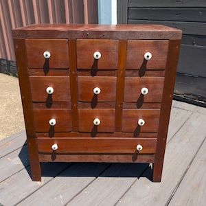 May include: A brown wooden chest of drawers featuring nine small square drawers arranged in three columns and one long horizontal drawer at the bottom. Each drawer is equipped with a small, round, white ceramic knob centered on the front panel.