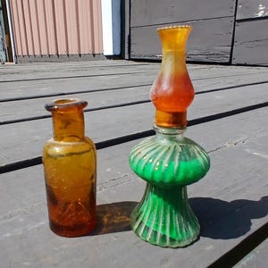 May include: Two antique glass bottles. One is amber-colored, cylindrical, and small. The other is a decorative lamp-shaped bottle with a green base, a clear center, and an orange top. Both bottles are on a wooden surface.