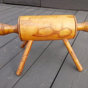 May include: A light brown wooden rolling pin with a cylindrical body and two handles on the ends, supported by three angled legs attached to the underside. The wood features a natural grain pattern with darker brown markings and decorative rings.