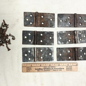 May include: An assortment of vintage metal hinges with visible rust, alongside a pile of screws. A wooden ruler with text is also present. The hinges have multiple pre-drilled holes for mounting.