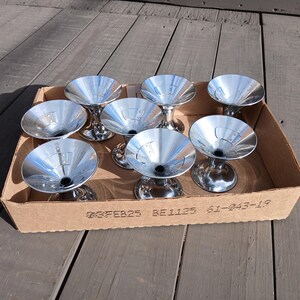 May include: A set of eight shiny, silver-colored martini glasses with a classic cone-shaped bowl and a short stem, presented in a cardboard box. The glasses are arranged neatly, reflecting the light. The box has printed text.