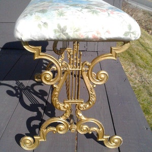 May include: A decorative bench with a gold-colored lyre-shaped base and a cushioned seat. The seat features a floral pattern with green leaves and pink flowers on a white background. The bench is designed for indoor use.
