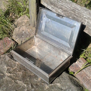 May include: An open, rectangular metal box with a hinged lid. The box is made of a dull silver-colored metal and sits on a rough stone surface. The lid is slightly ajar, revealing the empty interior.