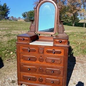 May include: A wooden dresser with a marble top, featuring four large drawers and two smaller upper drawers. An arched mirror with ornate carved wood detailing is attached to the top. Each drawer is equipped with a small, dark, round knob.