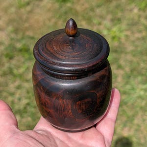 May include: A small, round, dark brown wooden jar with a matching lid featuring a prominent, pointed finial. The wood grain displays swirling, organic patterns in deep espresso and mahogany tones, creating a smooth, polished, and rounded vessel.