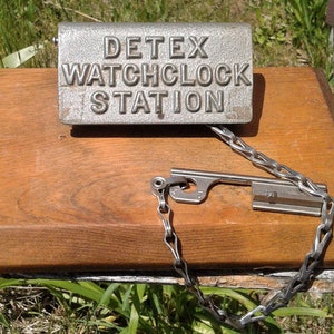 May include: A vintage Detex Watchclock Station sign made of metal, mounted on a wooden base. A metal key and chain are attached. The sign is weathered, suggesting age and use.