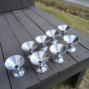 May include: A collection of eight shiny, silver-colored metal dessert cups. Each cup has a wide, shallow bowl and a pedestal base. The cups are arranged on a weathered, gray wooden surface.