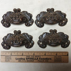 May include: Four antique bronze-colored drawer pulls with ornate floral designs. Each pull has two screw holes and a curved handle. A wooden ruler with text is below the pulls, measuring in inches.