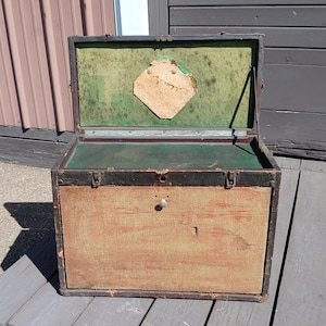 May include: An open rectangular storage trunk with a weathered tan exterior, dark brown metal trim, and two front latches. The interior is lined with green fabric and features a central diamond-shaped paper label on the inside of the lid.