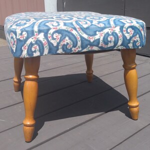 May include: A footstool with a cushioned top upholstered in a blue and white fabric featuring a swirling pattern with small pink floral accents. The piece is supported by four turned wooden legs with a smooth, light brown finish and tapered feet.