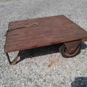 May include: A rustic, wooden cart with a rectangular platform and metal wheels. The cart has a dark brown, weathered appearance, with a rope attached to one side. The wheels are made of metal and have a worn, aged look.