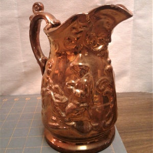 May include: A copper-colored pitcher with a detailed relief design. The pitcher features a handle and a spout, and the relief depicts figures in a pastoral scene. The pitcher has a glossy finish and is likely made of ceramic or metal.