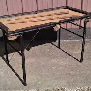 May include: A vintage, portable pool table with a black metal frame and tan felt playing surface. Two wooden cues rest on the table. The table has a folding leg design and small pockets. The table is approximately 48 inches long.