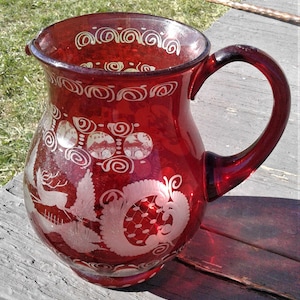 May include: A deep red glass pitcher featuring etched white designs of a deer, pine trees, and intricate floral patterns. The vessel has a wide mouth, a curved handle, and a series of decorative scrollwork motifs encircling the upper rim area.